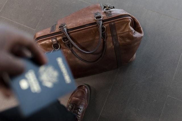person standing next to a brown leather duffel bag holding a passport